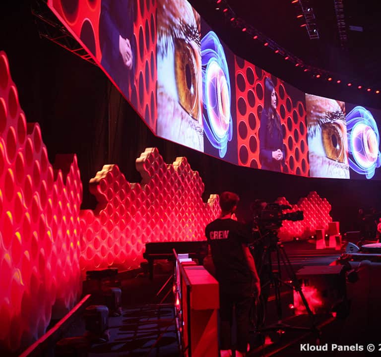 A stage with red honeycomb-patterned panels and large screens displaying eye images, with a crowd seated on the right. 