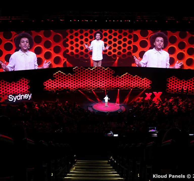 A person stands on stage delivering a presentation with red and white dot patterns on a large screen backdrop. The word 