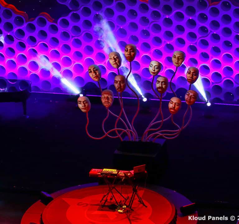 Stage setup with a honeycomb-patterned backdrop, spotlighted faces on elevated displays, a piano on the left, a keyboard on a circular red carpet, and an empty audience area.