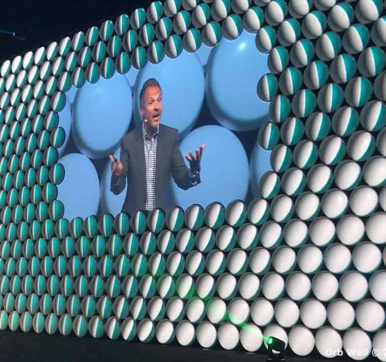 A man is speaking on stage, with his image projected on a large screen made of cylindrical shapes. The stage is lit with green and blue lights.
