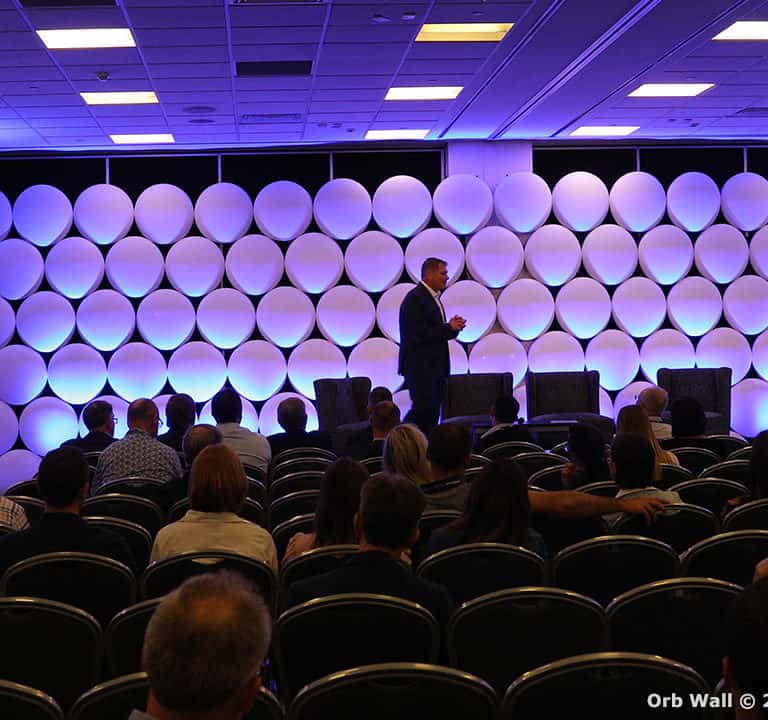 A speaker presents on stage in front of an audience. The backdrop consists of circular, illuminated panels. Several rows of seated attendees listen attentively.