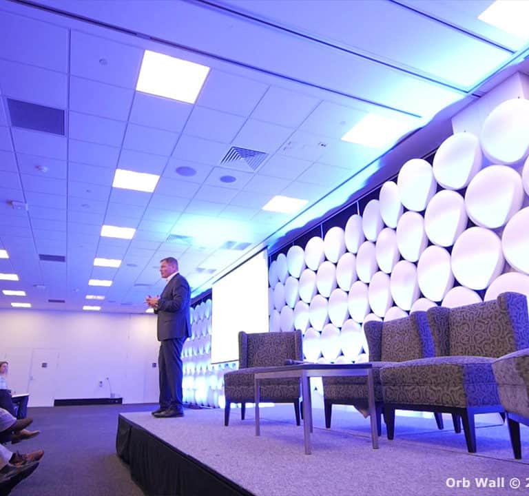 A speaker stands on a stage with a geometric backdrop and addresses an audience seated in front. Several chairs are arranged on the stage under bright, even lighting.