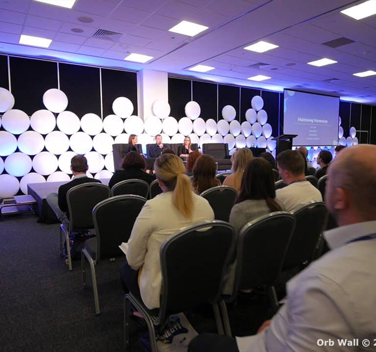 Audience seated and facing a stage where two presenters are speaking at a conference, with a backdrop of illuminated white circular patterns.