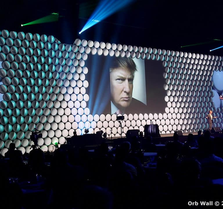 A speaker stands on a stage in front of a large, illuminated orb wall backdrop with projected images, addressing an audience in a darkened room. Text at bottom right reads