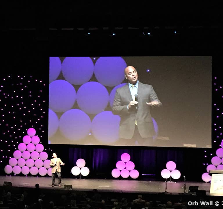 A man in a suit delivers a speech on a large stage with a modern, brightly lit backdrop consisting of circular light designs. The audience is seated in the foreground.