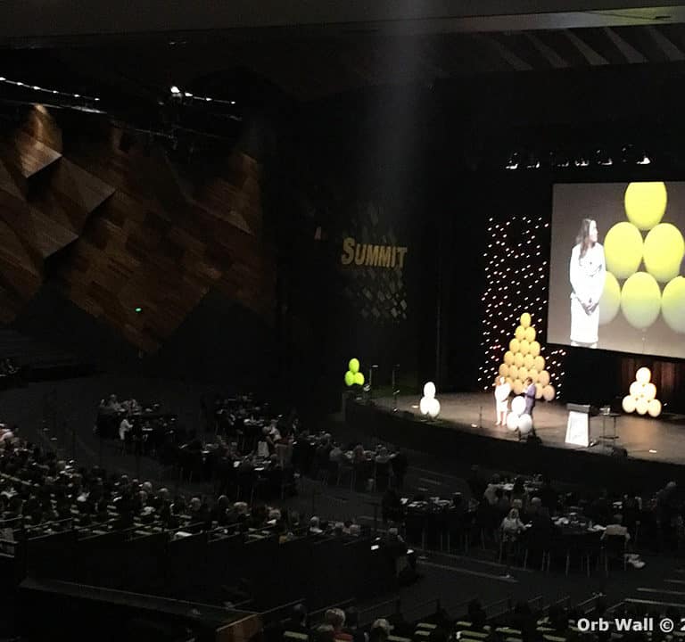 A large audience sits in an auditorium watching a presentation on a stage with speakers and large yellow balloons. The backdrop has the word 