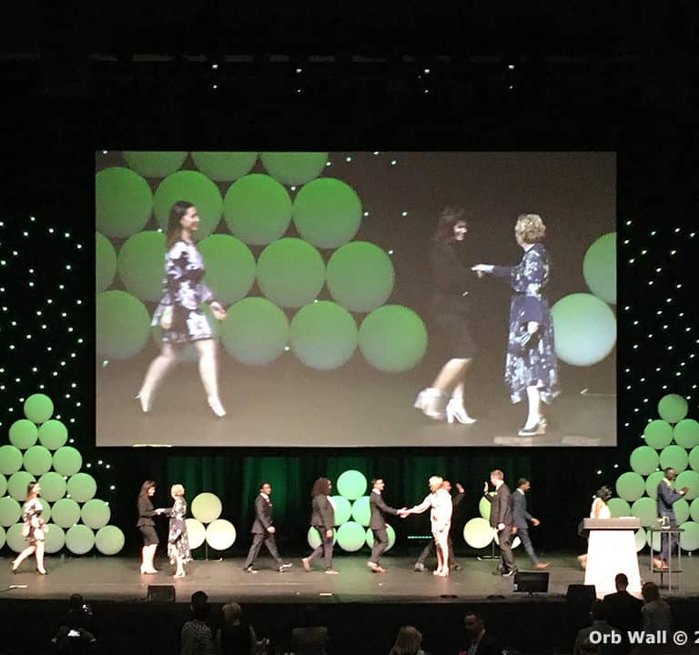 A group of people walking across a stage with a large screen displaying their movements. The backdrop features green circular decorations and star-like lights. Annotated 