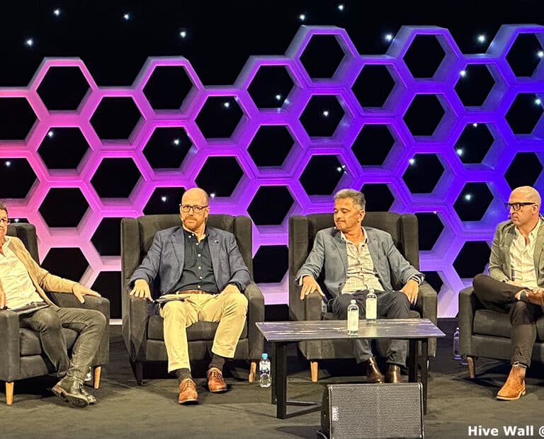 Four men seated on stage in front of a hive modular set, engaged in discussion.